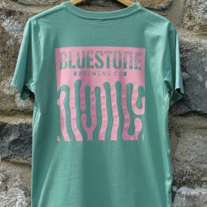 Green t-shirt with pink graphic design on a wooden hanger against a stone wall.