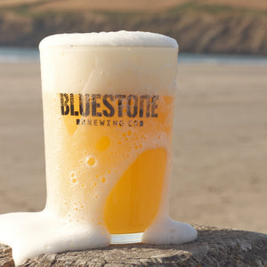 A frothy pint of Bluestone Brewing Co beer in a clear glass mug, sitting on a wooden post with a blurred beach and dunes in the background.