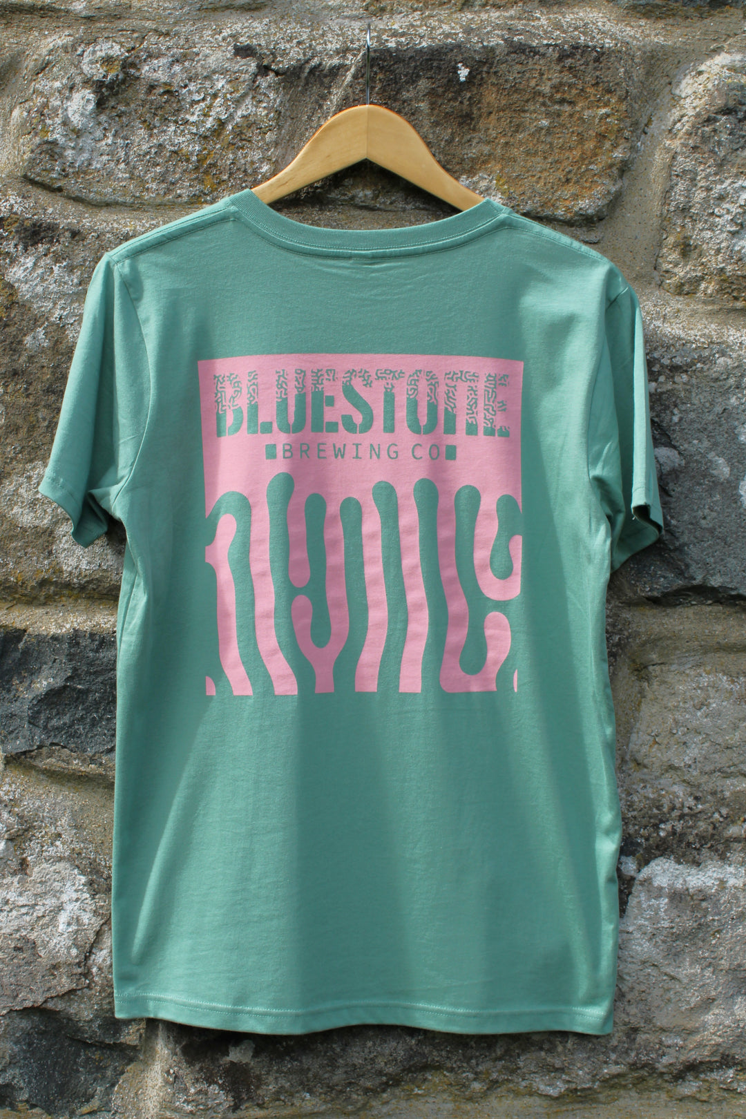 Green t-shirt with pink graphic design on a wooden hanger against a stone wall.